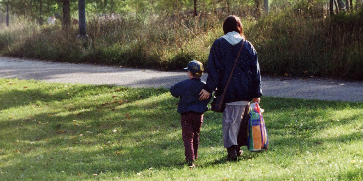 Kvinna och pojke som går i parkmiljö, sedda bakifrån. Kvinnan håller handen på pojken.