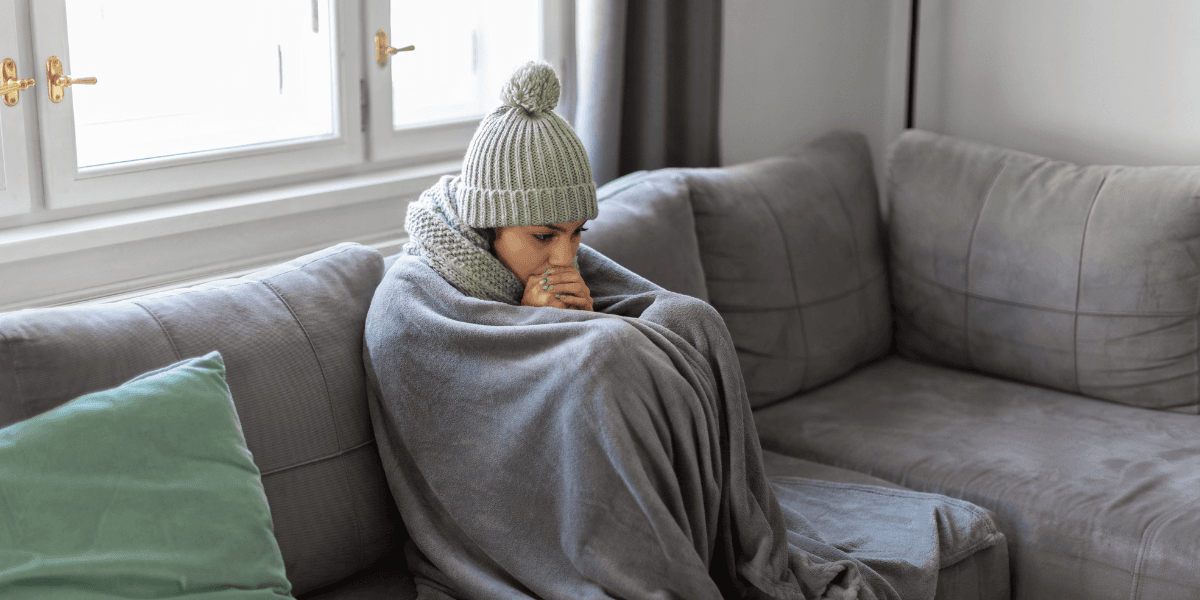 Person med mössa och filt sitter frusen på en soffa i ett kallt vardagsrum.