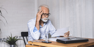 Äldre man granskar pensionsdokument vid köksbord, ser bekymrad ut.