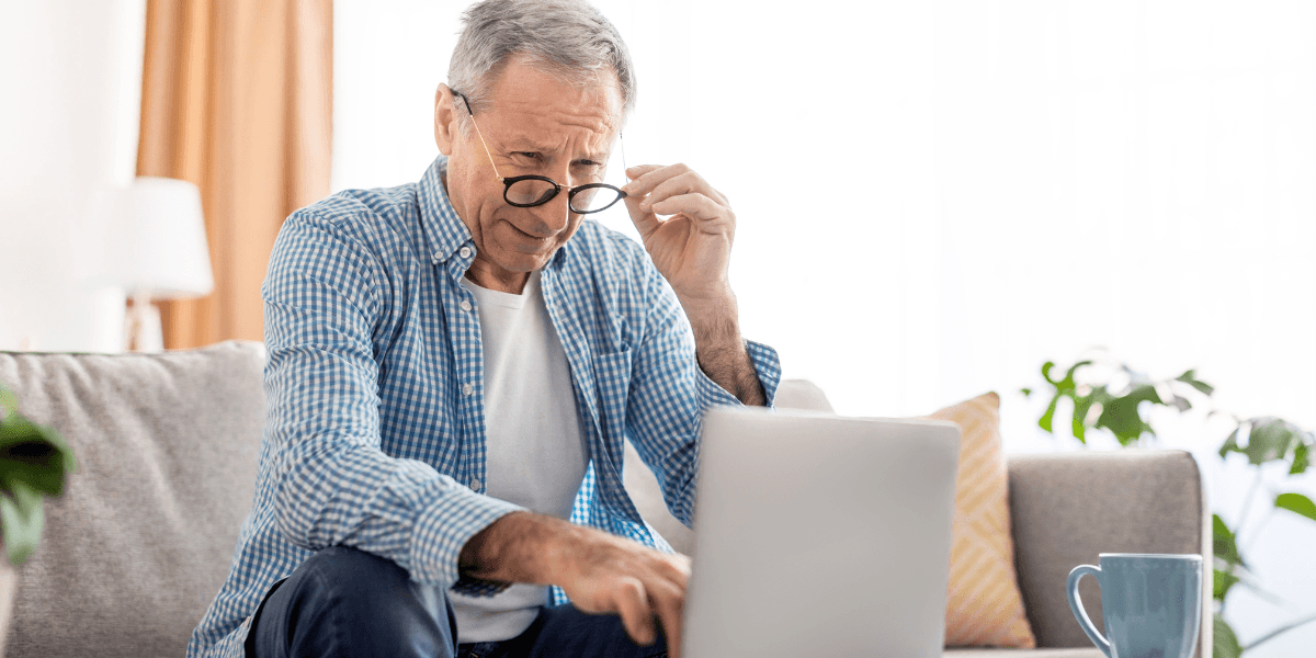Äldre man sitter i vardagsrum och tittar på laptop, symboliserar pensionär som kollar sin pensionsutbetalning online.