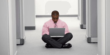 Anställd utan skrivbord sitter på golvet med dator – illustrerar platsbrist i kontorsmiljö.