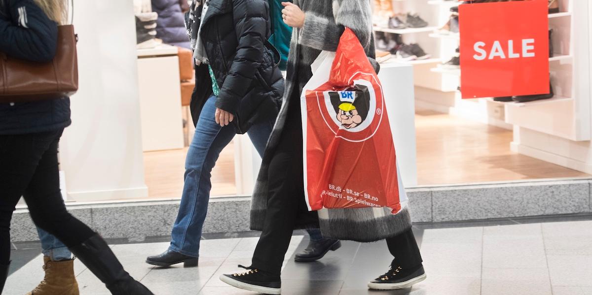 Personer går förbi en butik med "SALE"-skylt, en av dem bär en påse från en leksaksaffär.